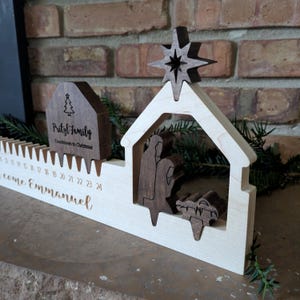 May include: Wooden Christmas countdown calendar featuring a nativity scene with a star, a house-shaped piece with the text "Pritzl Family Countdown to Christmas", and the words "come Emmanuel". The calendar has slots for each day.