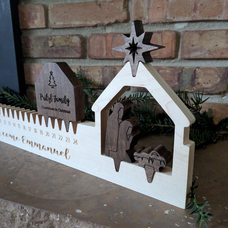 May include: Wooden Christmas countdown calendar featuring a nativity scene with a star, a house-shaped piece with the text "Pritzl Family Countdown to Christmas", and the words "come Emmanuel". The calendar has slots for each day.