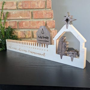 May include: Wooden Christmas countdown calendar with a nativity scene and star. The calendar has a countdown from 24 to 1, with the words "Oh come, oh come Emmanuel" engraved on the front. A small wooden house with the text "Pritzl Family" is also included.