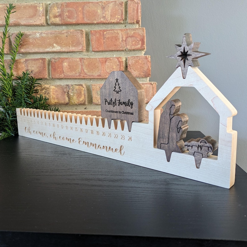 May include: Wooden Christmas countdown calendar with a nativity scene and star. The calendar has a countdown from 24 to 1, with the words "Oh come, oh come Emmanuel" engraved on the front. A small wooden house with the text "Pritzl Family" is also included.