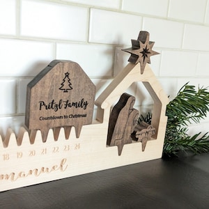 May include: Wooden Christmas countdown calendar with a nativity scene and a star. The calendar includes a small house with the text "Pritzl Family Countdown to Christmas" and a tree graphic. The numbers 18-24 are visible.