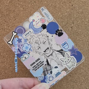May include: A clear, rectangular charm with a black and white anime-style illustration and various stickers. The stickers include a bear, paw prints, a bunny, and the text "COOL GIRL". A blue beaded chain hangs from the top.