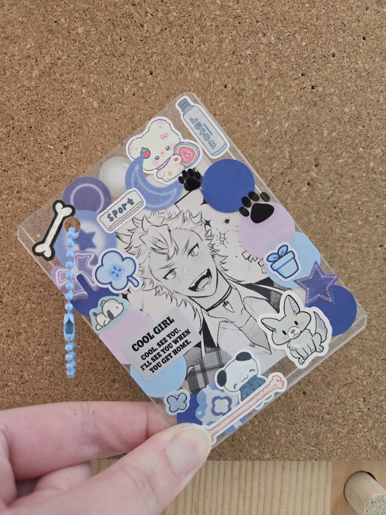 May include: A clear, rectangular charm with a black and white anime-style illustration and various stickers. The stickers include a bear, paw prints, a bunny, and the text "COOL GIRL". A blue beaded chain hangs from the top.