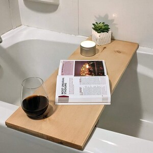 May include: A wooden bath tray with a book, a glass of wine, and a candle. The tray is placed on the edge of a white bathtub.