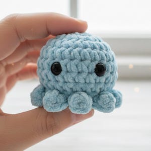 Könnte beinhalten: Ein kleiner, hellblauer gehäkelter Oktopus mit schwarzen Sicherheitsaugen. Das Plüschtier hat einen runden Kopf und acht kurze, abgerundete Tentakel. Der weiche, handgefertigte Oktopus wird in einer Hand gehalten.