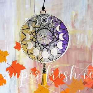 May include: A purple and gold circular wall hanging with a silver tassel. The hanging features a silver wire design with a circle in the centre that says "Aries". The circle is surrounded by silver crescent moons.