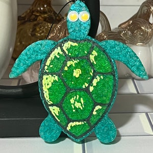 May include: A teal blue sea turtle ornament with a green shell and yellow accents. The turtle has white googly eyes.