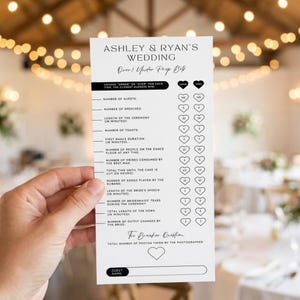 May include: A white wedding game card with black text, held by a hand. The card lists categories like "Number of Guests" and "Number of Speeches" for guests to guess. The card is titled "Ashley & Ryan's Wedding".