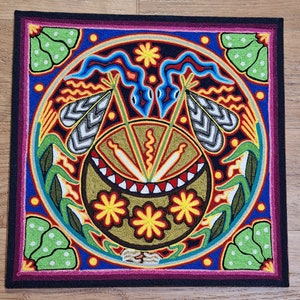 May include: A square, embroidered textile with a central circular design. The design features a gold-colored bowl with feathers, surrounded by blue, red, yellow, and green patterns. The border is black, with a purple and green outer edge.