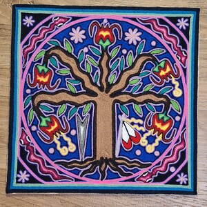 May include: Square textile art piece featuring a central brown tree with green leaves and red and yellow flowers. The tree is surrounded by a blue background with a pink circle and a black border. The design includes floral and geometric elements.