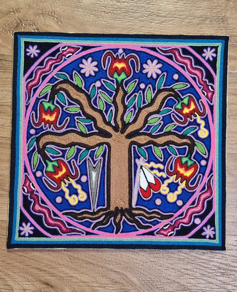 May include: Square textile art piece featuring a central brown tree with green leaves and red and yellow flowers. The tree is surrounded by a blue background with a pink circle and a black border. The design includes floral and geometric elements.