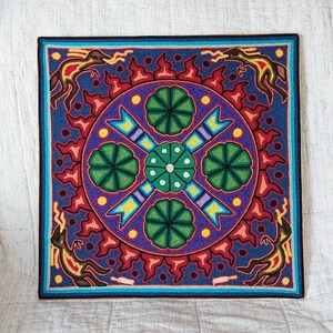 May include: Square, colorful wall art with a detailed, symmetrical design. The artwork features a central circular pattern with green floral elements, surrounded by a blue background and a border with stylized figures and red accents. The piece is likely handmade.