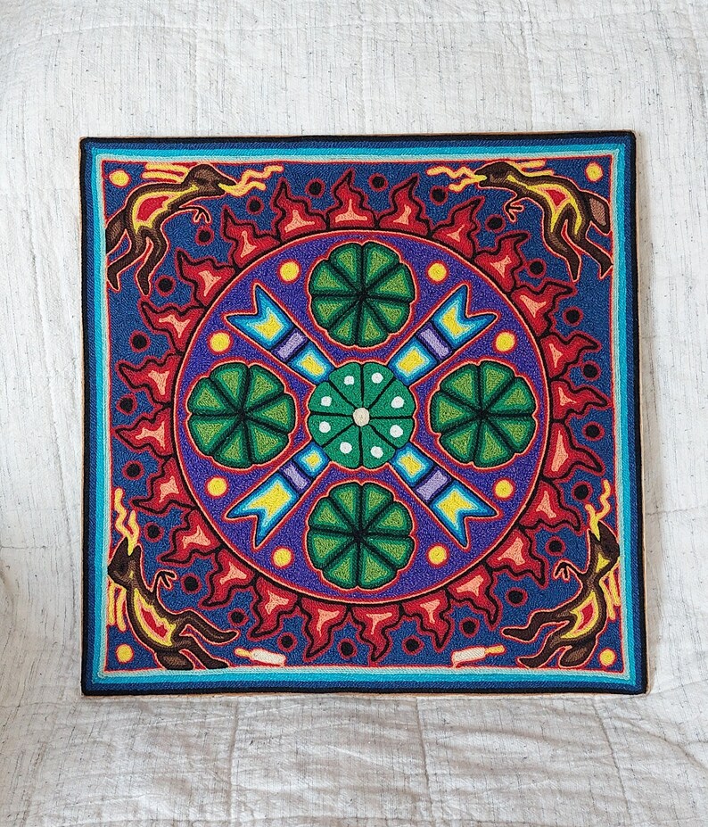 May include: Square, colorful wall art with a detailed, symmetrical design. The artwork features a central circular pattern with green floral elements, surrounded by a blue background and a border with stylized figures and red accents. The piece is likely handmade.