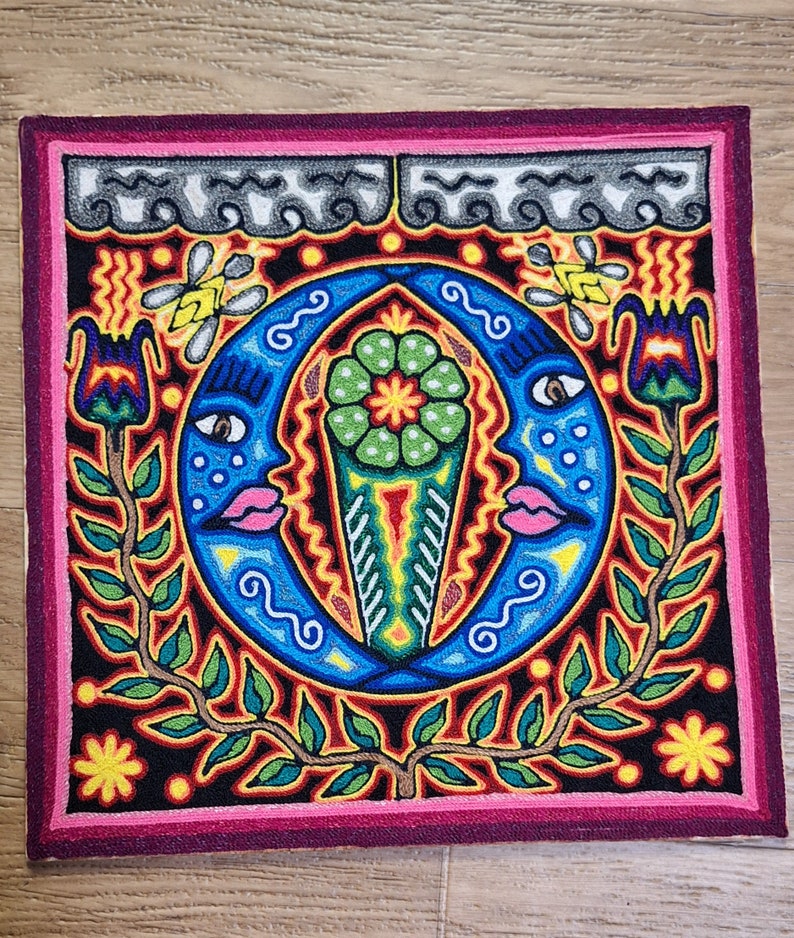 May include: A colorful embroidered artwork with a black background. The artwork features two blue crescent moons with faces, a green flower, and a vine with green leaves and pink flowers. The artwork is framed with a pink border.