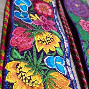 May include: A close-up of a colorful embroidered fabric with a floral pattern. The fabric is purple with yellow, orange, red, blue, and green flowers. The flowers are embroidered with intricate details.