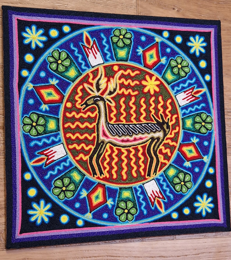 May include: Square, beaded artwork featuring a central deer figure with antlers, surrounded by a circular design. The artwork uses vibrant colors like blue, red, yellow, and green. The deer is outlined in black, with a pink and white body. The border is black, with a pink and purple trim.