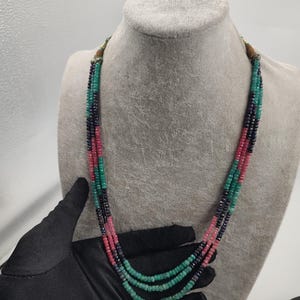 以下が含まれることがあります： 小さなカラフルなビーズが連なった、マルチストランドネックレス。緑、ピンク、濃い青色のビーズが組み合わされています。グレーのマネキンの頭に飾られています。
