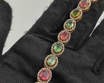 May include: A gold bracelet with oval-shaped gemstones in various colors, including pink, green, and orange. The gemstones are surrounded by smaller, clear gemstones.
