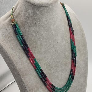 Könnte beinhalten: Eine mehrsträngige Halskette mit abwechselnden Abschnitten aus grünen, roten und schwarzen Perlen. Die Halskette wird auf einer Mannequin-Büste präsentiert. Die Perlen sind klein und rund und erzeugen einen geschichteten Effekt. Die Halskette hat einen dekorativen Verschluss.