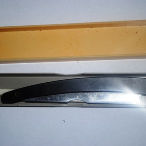 May include: A straight razor with a black handle and a silver blade, resting in a clear plastic case. The case is inside a light orange rectangular box. The razor is designed for shaving.