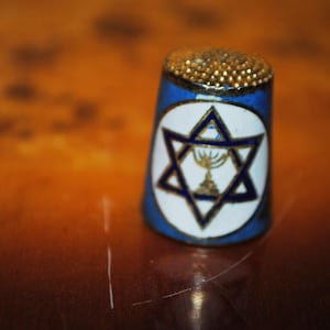 Op de afbeelding: Een blauwe vingerhoed met een gouden kralen bovenkant en een witte ovaal met een Davidster en een menora. De vingerhoed is een naai-instrument, waarschijnlijk voor naaiwerk of knutselen.