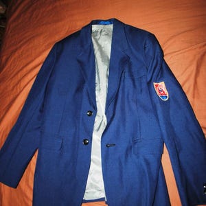 May include: A navy blue blazer with a light grey interior lining. The blazer has three black buttons and a small patch on the left sleeve. The blazer is lying on a brown surface.
