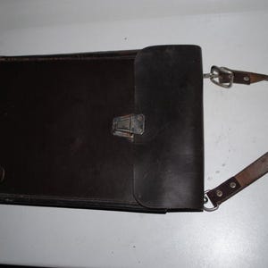 May include: A black leather portfolio with a flap closure and a brown leather shoulder strap. The portfolio has a metal clasp and a metal buckle on the strap. The item is likely vintage.