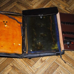 May include: A brown leather case with a flap that opens to reveal a clear plastic window and a compartment with multiple slots. The case has a metal clasp and a strap.