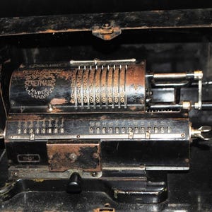 Op de afbeelding: Een antieke mechanische rekenmachine van zwart en roestig metaal, open in de koffer. De rekenmachine heeft meerdere nummerwielen en hendels. De merknaam "SCHEUTZ" is zichtbaar aan de zijkant.