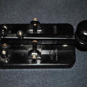 May include: A black telegraph key with a rectangular base, a horizontal metal arm, and a round knob. The device features metallic contact points, adjustment screws, and a small embossed logo on the surface of the base, all finished in a glossy black.