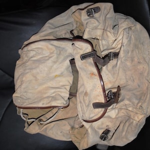 May include: A vintage canvas backpack in a faded beige color, featuring multiple compartments and leather straps with metal buckles. The bag shows signs of wear, suggesting it's a well-used item. The design is reminiscent of military or outdoor gear.