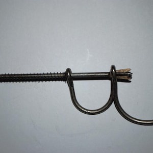 May include: A vintage metal corkscrew with a long, threaded shaft and a circular handle. The design includes a curved handle and a small, circular plate at the end of the shaft. The corkscrew is designed to remove corks from bottles.