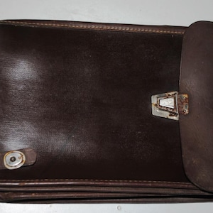 May include: A dark brown leather portfolio with a flap closure and a metal clasp. The portfolio is rectangular and appears to be vintage, with visible wear and tear. The flap is rolled over to the right side.