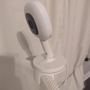 May include: A white baby monitor with a round camera lens mounted on a white stand. The stand is attached to a white wicker basket.