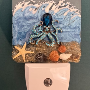 May include: Decorative night light featuring an ocean scene with a blue octopus, starfish, and seashells. The octopus has turquoise accents and a large blue gem. The background depicts ocean waves. The night light is white.