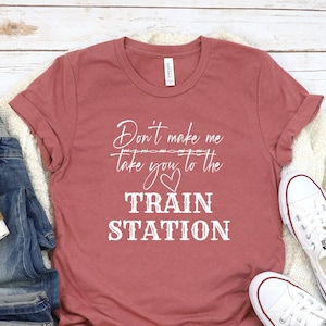 May include: A heather mauve t-shirt with white text that reads Don't make me take you to the TRAIN STATION with a heart graphic. The shirt is displayed with blue denim jeans, a white coffee cup, and white canvas sneakers on a white wood surface.