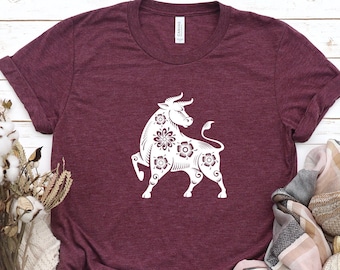 May include: A heather maroon t-shirt with a white graphic of a stylized bull with floral details. The bull is facing right and has its head turned to the left. The t-shirt is laying flat on a white wooden surface.
