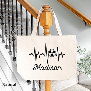 May include: A natural canvas tote bag with a black and white design of a heartbeat with a radiation symbol in the middle and the name "Madison" underneath.