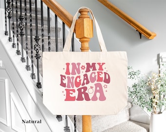 May include: A natural canvas tote bag with the text "In My Engaged Era" in pink and red letters with a diamond ring graphic.