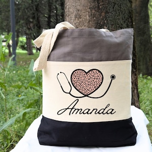 May include: A gray and white canvas tote bag with a black bottom. The bag has a heart with a leopard print design and a stethoscope. The name "Amanda" is printed below the heart.