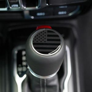 May include: A black gear shift knob with a black and gray American flag design. The shift knob is in a vehicle's interior, with other controls visible in the background.