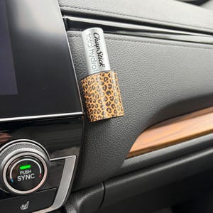 May include: A ChapStick total hydration lip balm with a leopard print sleeve is shown. The lip balm is silver with black text. The sleeve is tan with brown spots. The lip balm is attached to a car's dashboard.