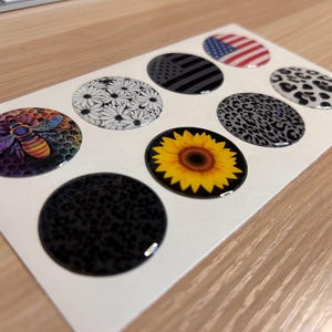 May include: A sheet of eight oval-shaped stickers with various designs. The designs include a bee, daisies, an American flag, a black and gray flag, leopard print, and a sunflower. The stickers are glossy and have a raised, three-dimensional appearance.