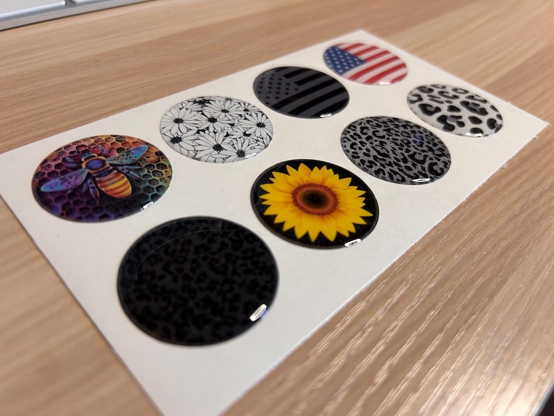 May include: A sheet of eight oval-shaped stickers with various designs. The designs include a bee, daisies, an American flag, a black and gray flag, leopard print, and a sunflower. The stickers are glossy and have a raised, three-dimensional appearance.