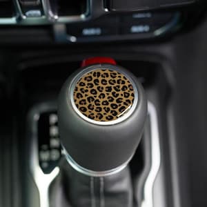 May include: A black gear shift knob with a leopard print design. The leopard print is a pattern of brown spots on a tan background. The knob is in a car interior.