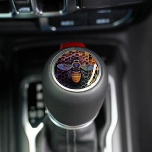 May include: Black gear shift knob with a colorful bee design. The bee is centered on a honeycomb background with shades of orange, yellow, and purple. The knob is in a vehicle's interior.