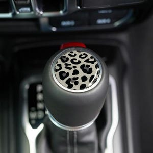 May include: A black gear shift knob with a silver trim and a leopard print design. The leopard print features black spots on a light gray background. The gear shift is in a vehicle's interior.