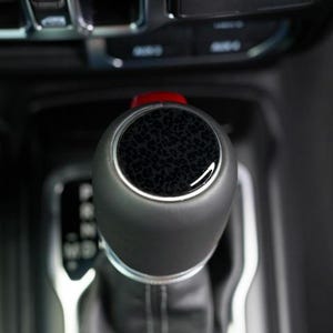 May include: Close-up of a black gear shift knob with a textured, dark pattern on top. The knob has a silver trim and a red accent at the base. The background is blurred, showing the interior of a vehicle.