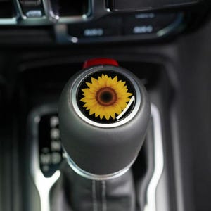 May include: A black gear shift knob with a sunflower design. The sunflower has yellow petals and a brown center. The knob is set against a dark interior background.