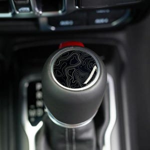 May include: A black gear shift knob with a topographic map design. The knob has a black, rounded shape with a silver trim. The top of the knob features a black and white map pattern. The shift lever is in a vehicle's interior.